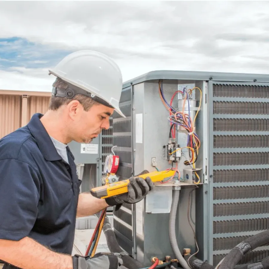 HVAC technician in a hard hat testing an outdoor air conditioner with a multimeter